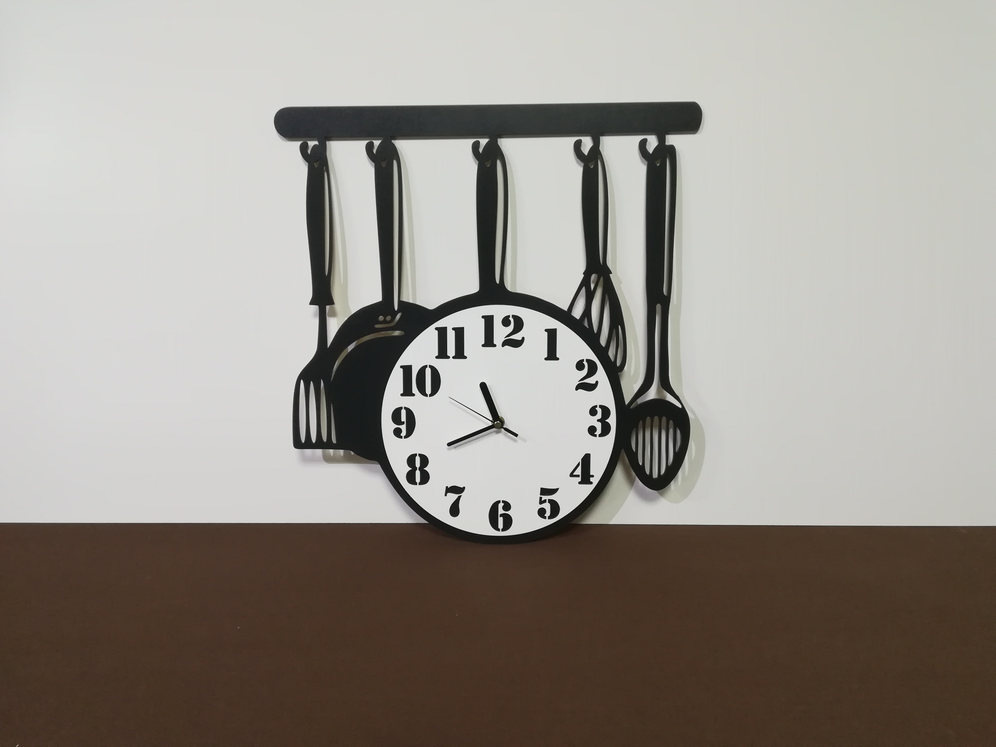 Dekoracyjny zegar ścienny w kształcie patelni, z zawieszonymi czarnymi przyborami kuchennymi na białej ścianie z brązowym pasem na dole.