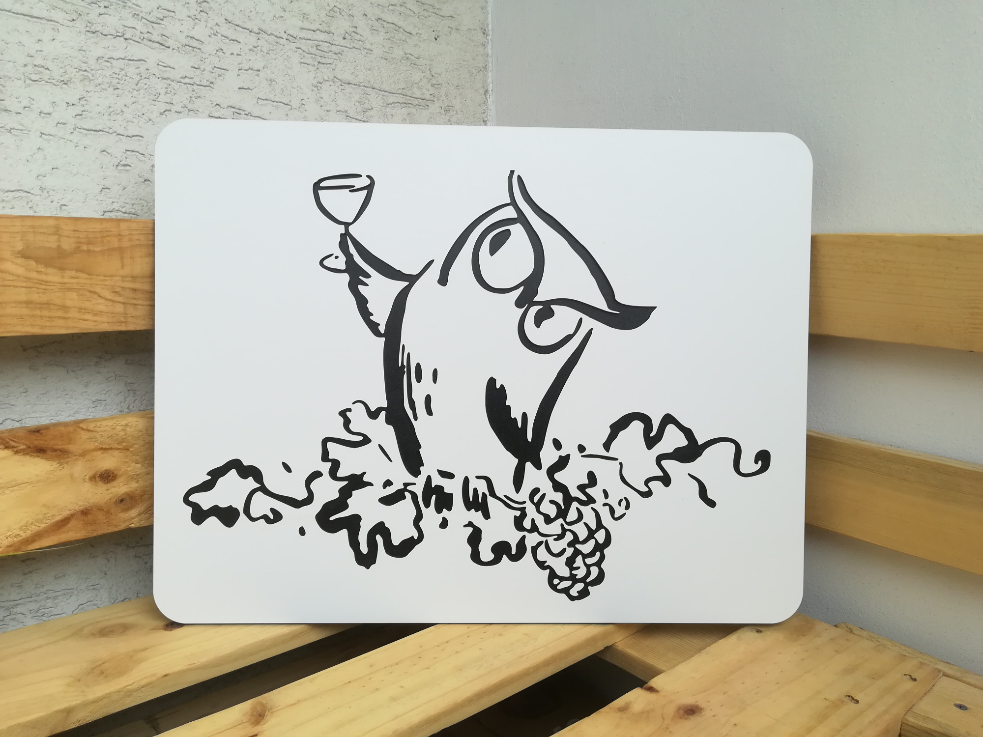 Ręcznie rysowana ilustracja sowy trzymającej kieliszek wina, umieszczona na białej tablicy z zaokrąglonymi rogami, leżącej na drewnianej palecie.