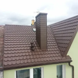 Brązowy dach z blachodachówki, dopasowany kolorystycznie komin z anteną, fragment elewacji w kolorze pistacjowym, widok z góry.