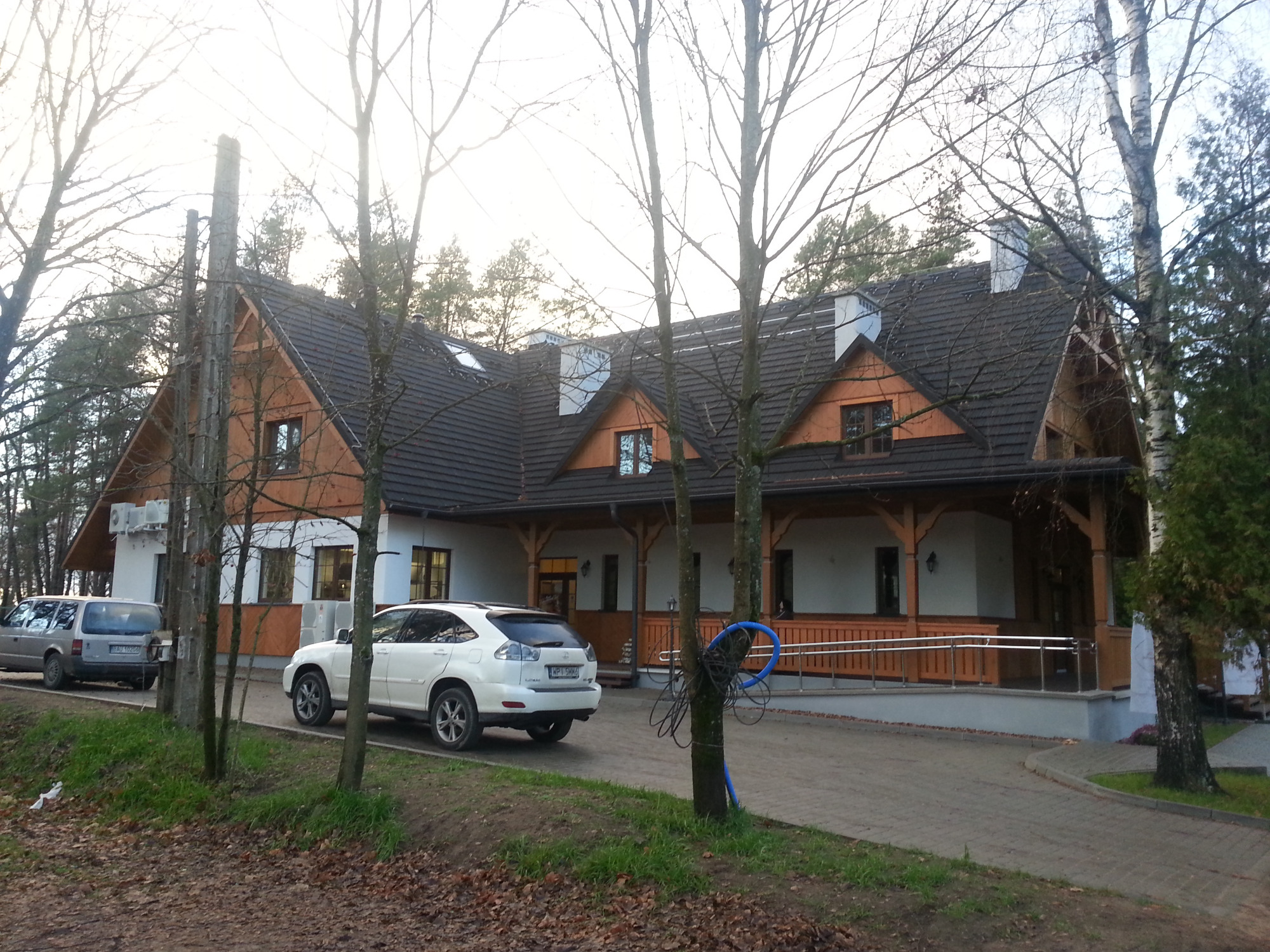 Dom z ciemnym dachem z dachówki ceramicznej, drewnianą więźbą dachową i białymi ścianami, widziany z zewnątrz przez drzewa bez liści. Na podjeździe zaparkowany biały samochód.