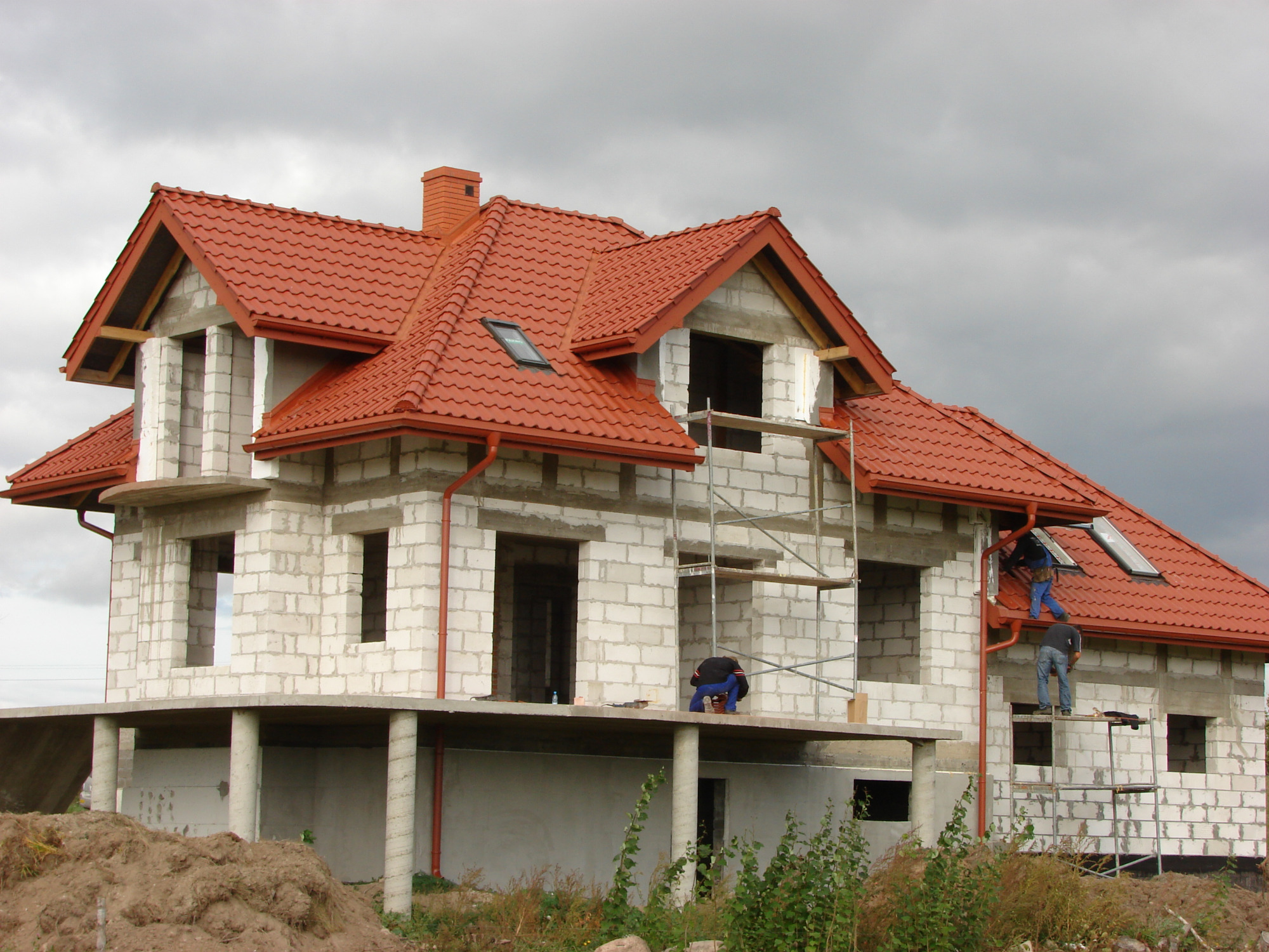 Dom w trakcie budowy z zamontowaną czerwoną dachówką i rynnami, pracownicy na rusztowaniu montują rynny, widoczna surowa konstrukcja budynku z betonu komórkowego.