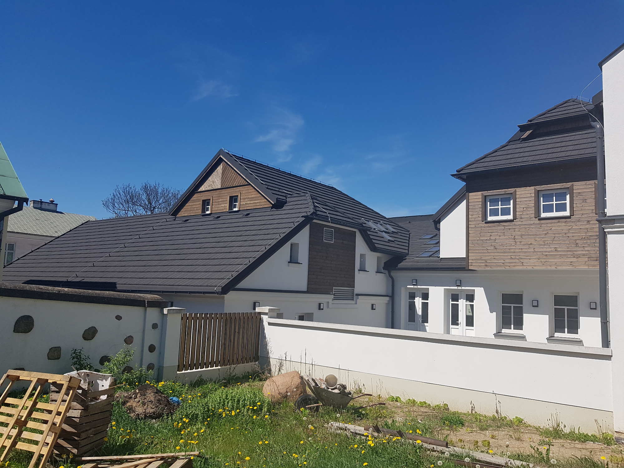 Nowoczesny dom z ciemną dachówką, drewnianą elewacją i białymi ścianami, widziany w słoneczny dzień z poziomu gruntu, z widocznymi paletami i taczką na pierwszym planie.