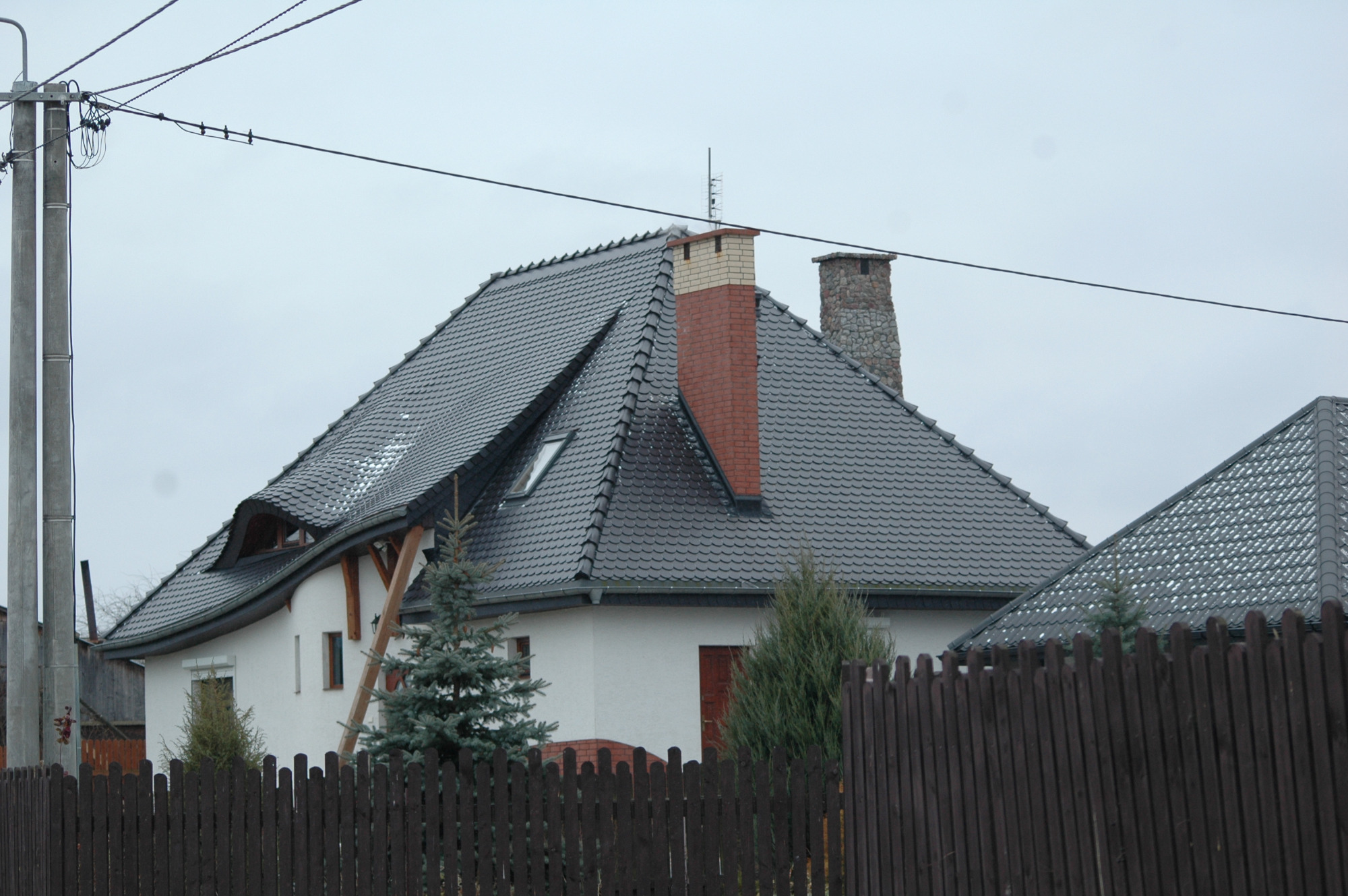 Dom jednorodzinny z ciemnoszarym dachem pokrytym dachówką karpiówką, widoczny komin z cegły klinkierowej, drewniana konstrukcja nad lukarną, i brązowe ogrodzenie sztachetowe na pierwszym planie.