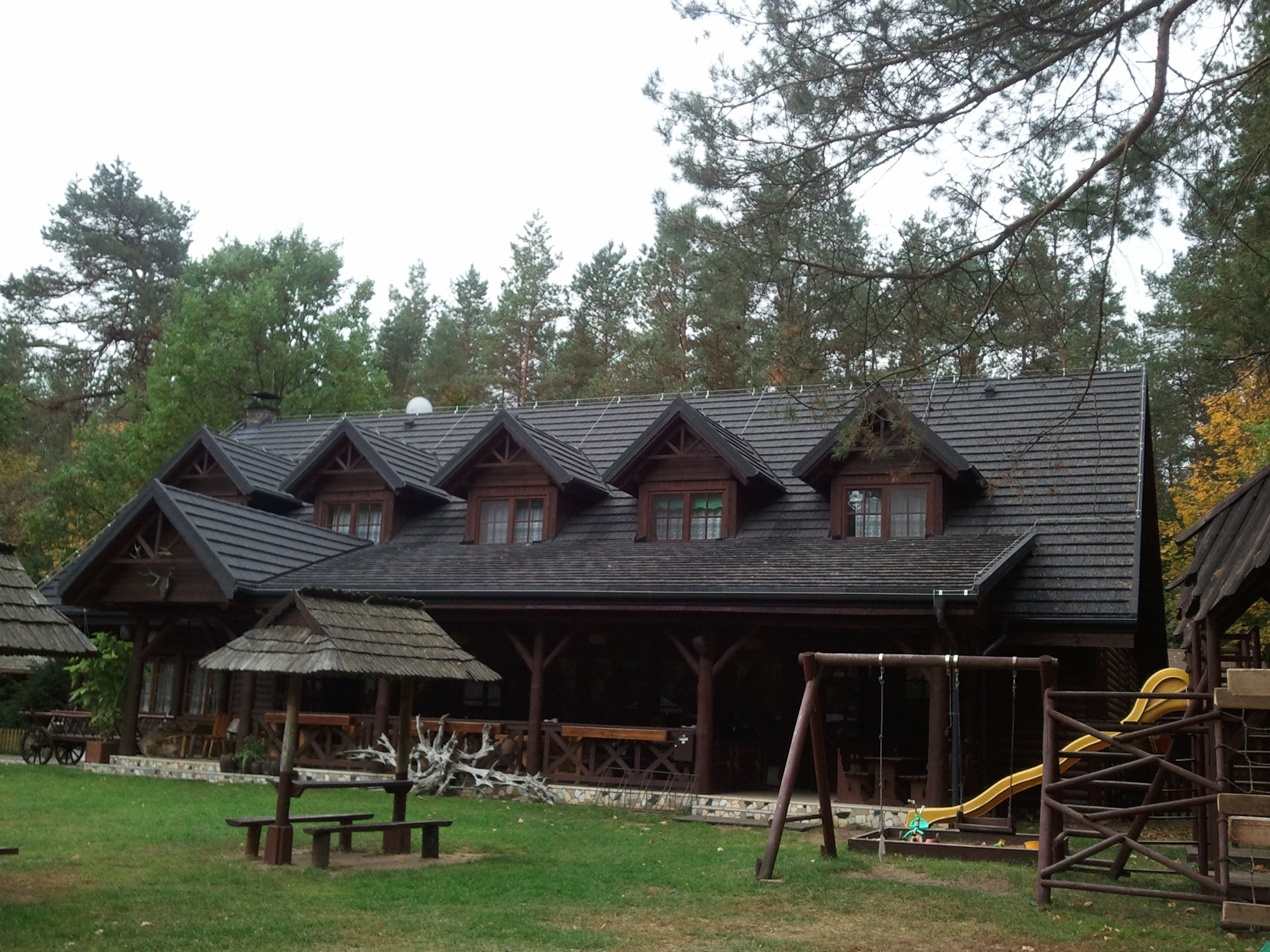 Drewniany budynek w stylu góralskim z ciemnym dachem krytym dachówką bitumiczną, widoczny z placu zabaw z huśtawkami i zjeżdżalnią.