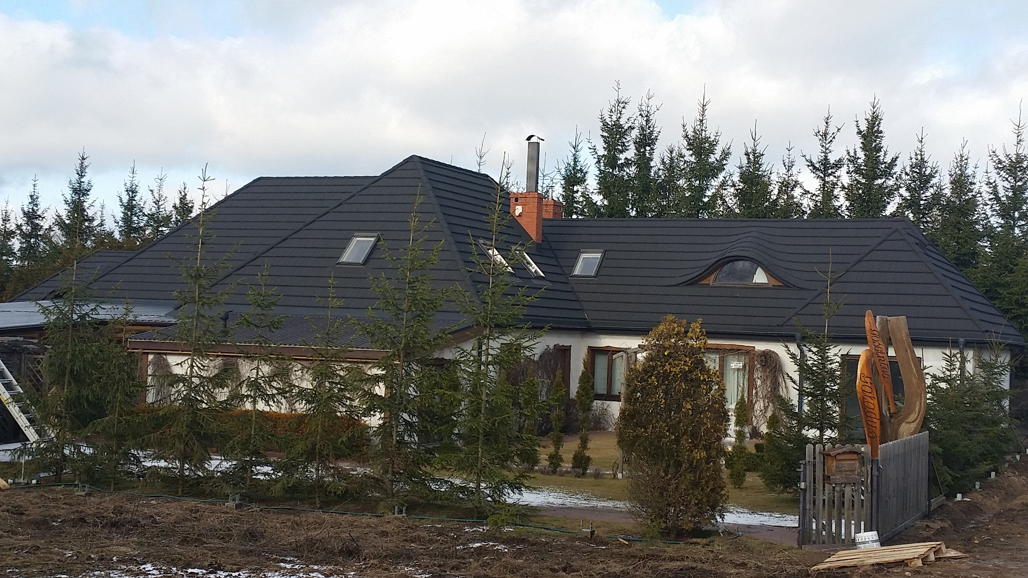 Dom jednorodzinny z ciemnoszarym dachem z dachówki bitumicznej, oknami dachowymi i kominem z cegły, otoczony iglakami i drewnianym ogrodzeniem z rzeźbioną dekoracją.