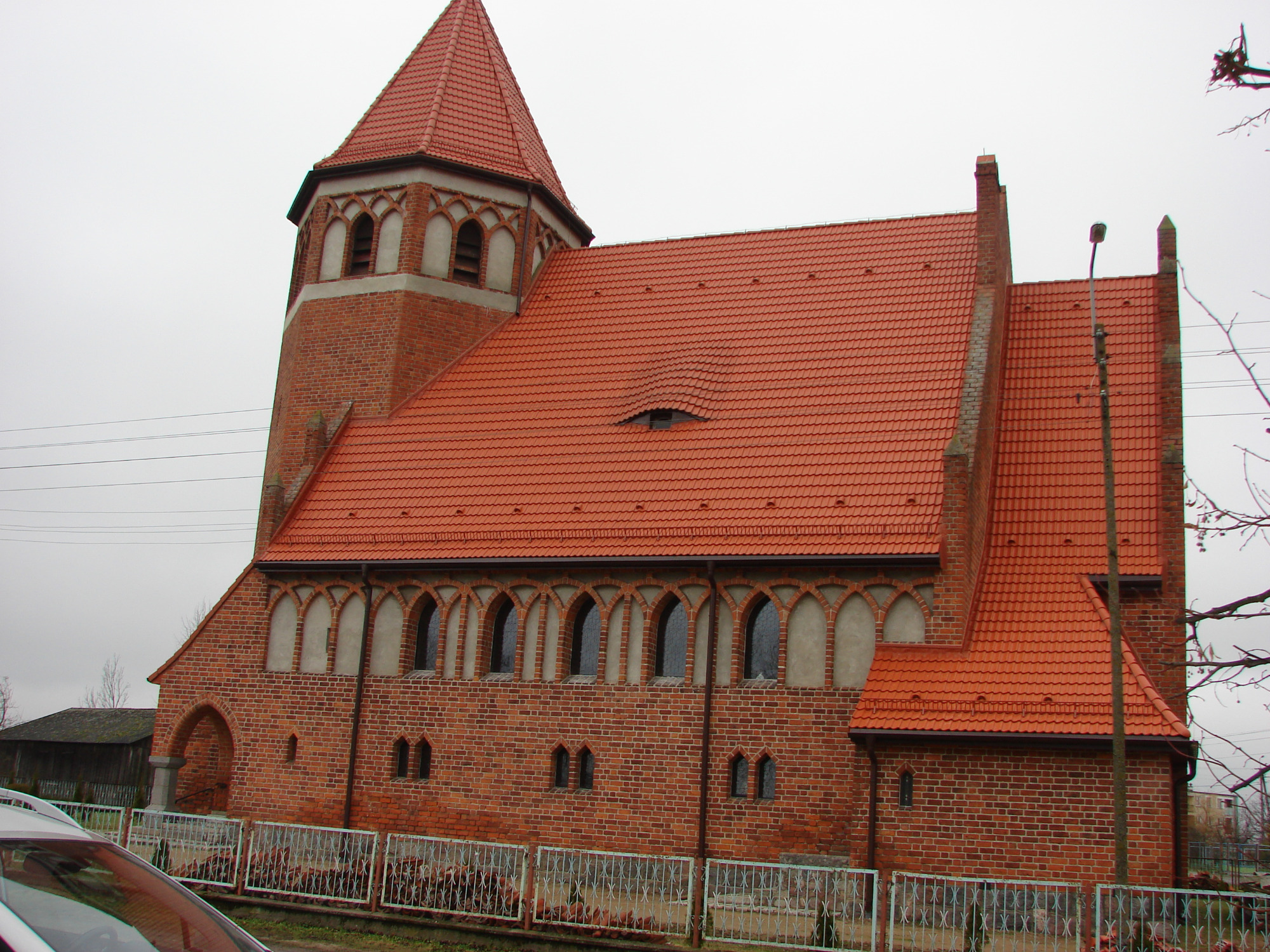 Czerwony ceglany kościół z nową, czerwoną dachówką ceramiczną, widoczny zza metalowego ogrodzenia.