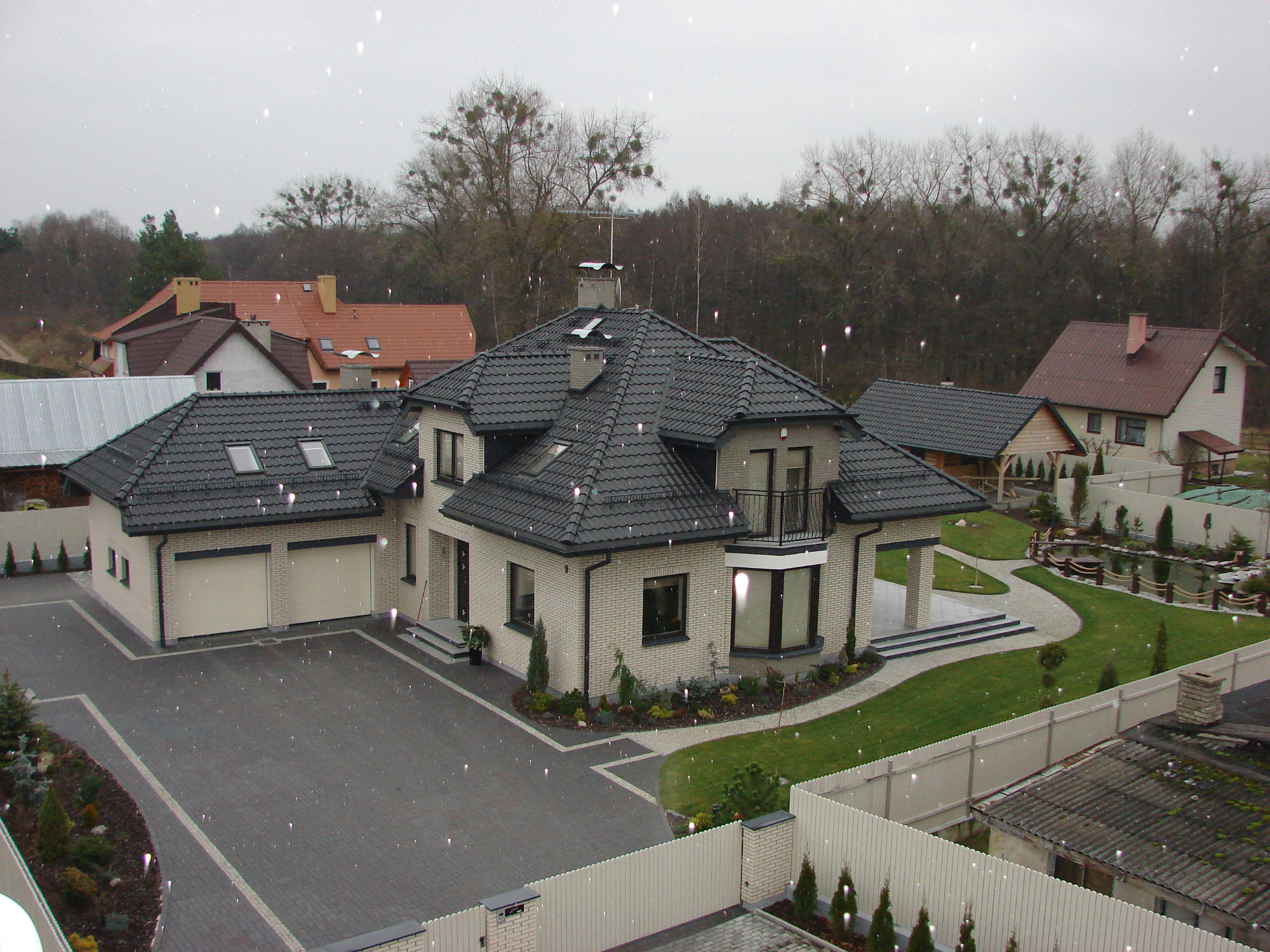 Nowoczesny dom z szarą dachówką karpiówką, widok z góry podczas opadów śniegu, z zadbanym ogrodem i oczkiem wodnym w tle.