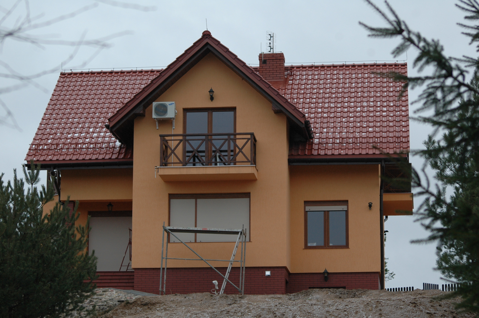 Dwukondygnacyjny dom z pomarańczową elewacją i czerwoną dachówką, z balkonem i klimatyzatorem na ścianie, częściowo zasłonięty przez drzewa iglaste, ze śladami śniegu na dachu.
