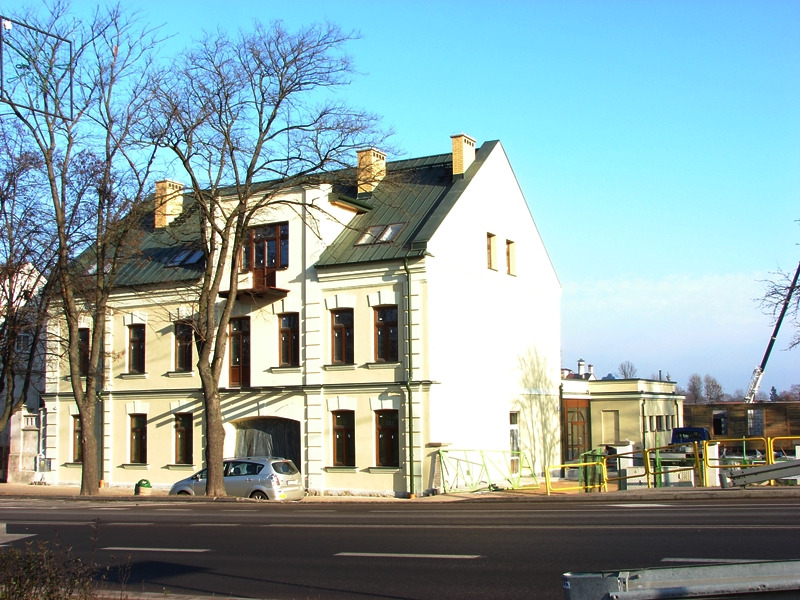 Odnowiona kamienica z zielonym dachem, widoczna z perspektywy ulicy, z samochodem zaparkowanym przed budynkiem i konstrukcją budowlaną w tle.
