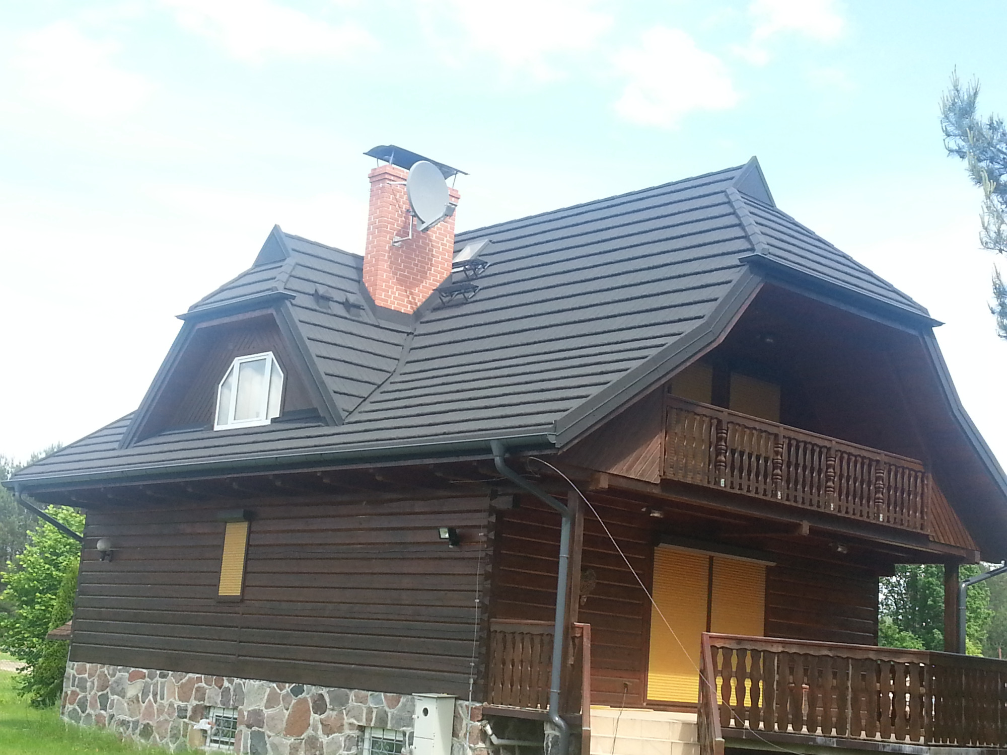 Dom z ciemnobrązową drewnianą elewacją, kamienną podmurówką i dachem z blachodachówki z kominem z anteną satelitarną.