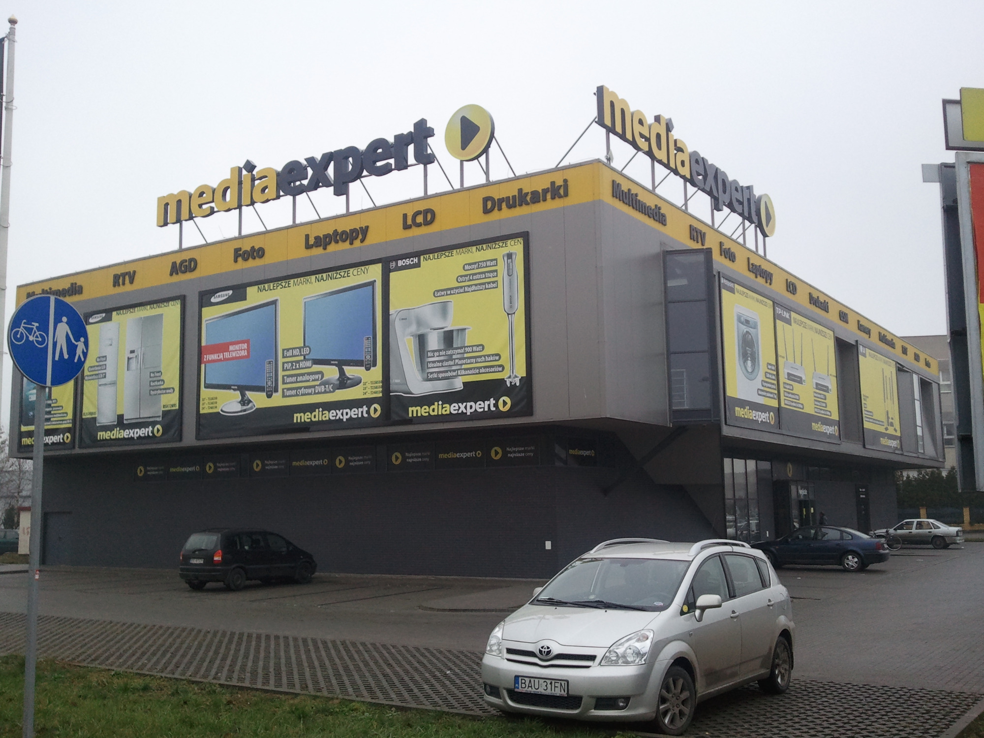 Budynek sklepu Media Expert z reklamami AGD, RTV i elektroniki, widok z parkingu z zaparkowanymi samochodami.