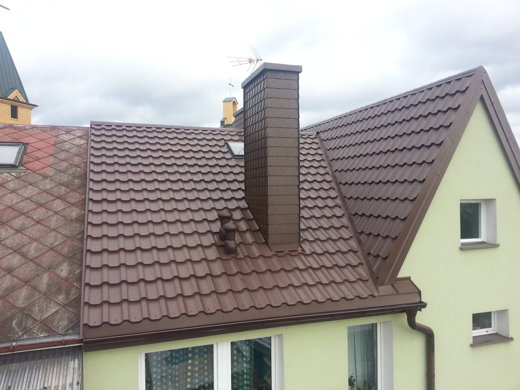 Brązowy dach z blachodachówki, dopasowany kolorystycznie komin z anteną, fragment elewacji w kolorze pistacjowym, widok z góry.
