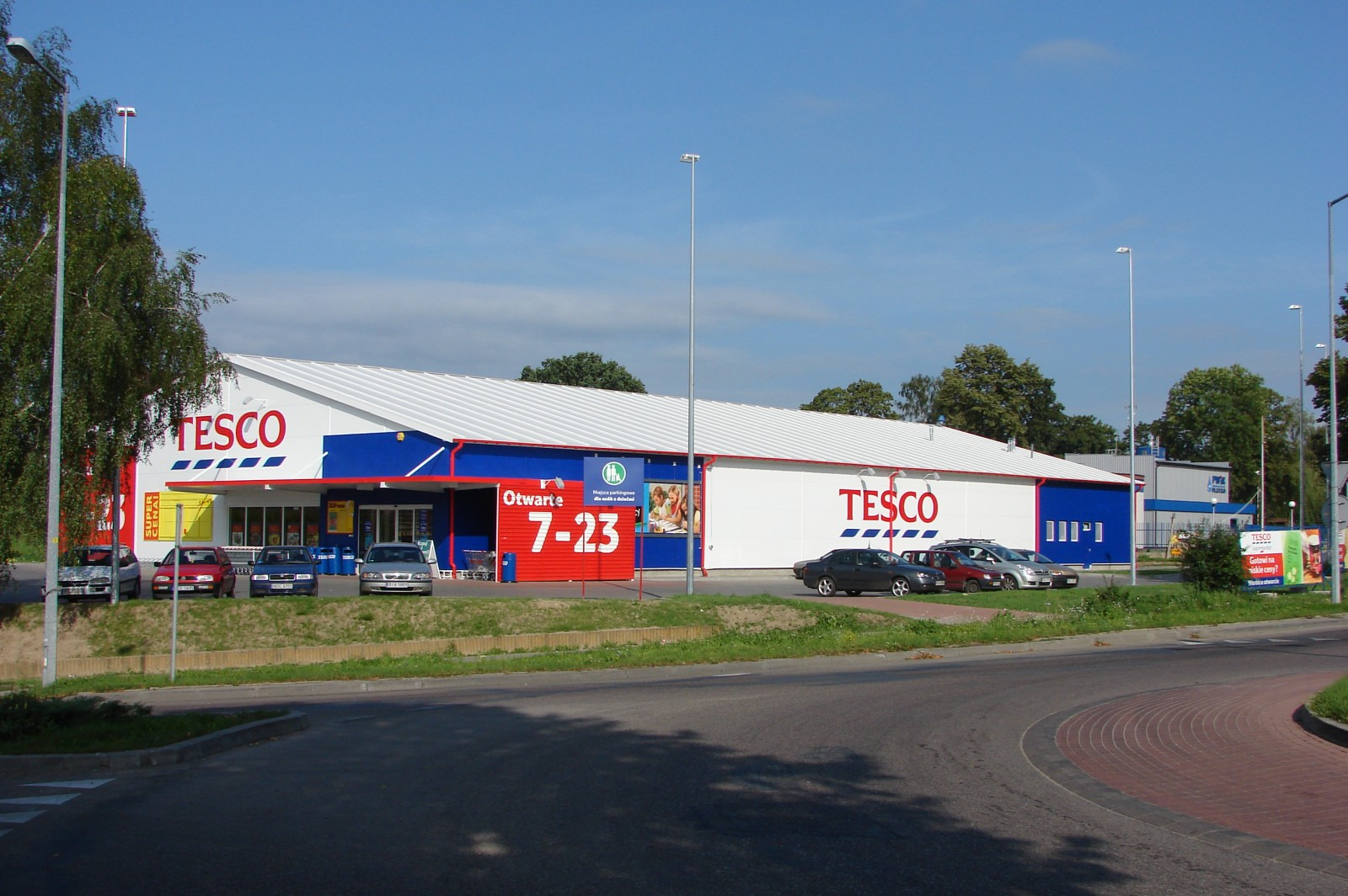 Duży budynek sklepu Tesco z białym dachem, niebieskimi akcentami i czerwonymi elementami reklamowymi, widoczny z ulicy z zaparkowanymi samochodami.