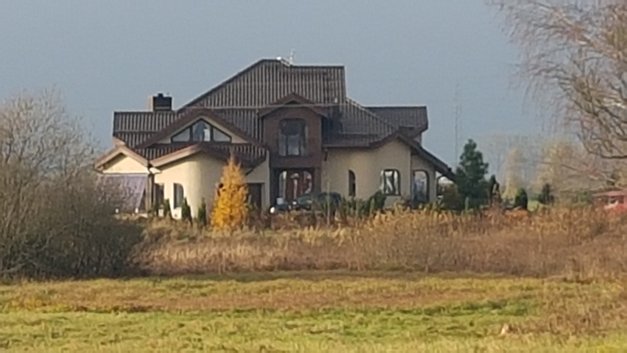 Dwupiętrowy dom jednorodzinny z brązowym dachem wielospadowym, widoczny z daleka na tle jesiennego krajobrazu z drzewami bez liści i suchą trawą.