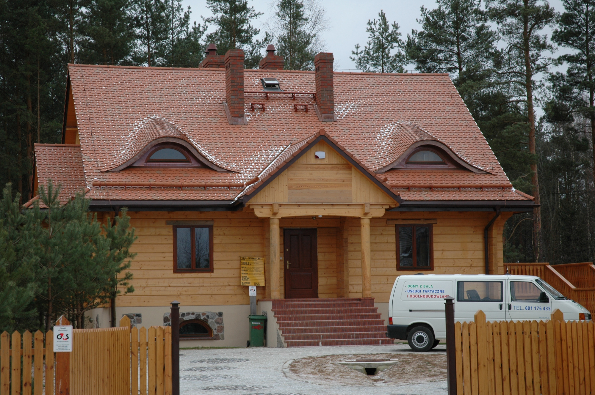 Dom z bali kryty dachówką karpiówką, z charakterystycznymi oknami w kształcie kocich oczu, widoczny z przodu, na tle lasu iglastego, z zaparkowanym samochodem dostawczym firmy budowlanej.