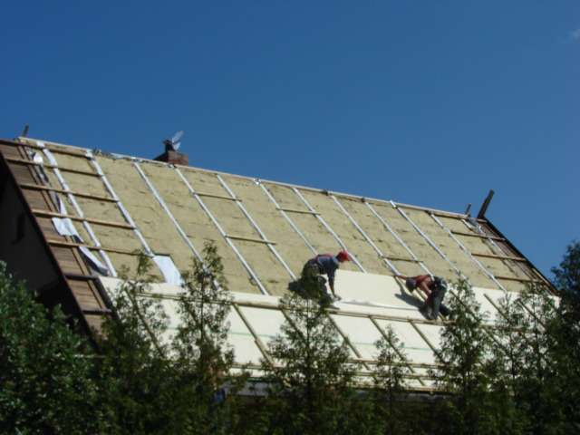 Montaż izolacji termicznej na dachu skośnym z widoczną więźbą, pracownicy w uprzężach zabezpieczających, fragment komina z wiatraczkiem i zieleń w tle.