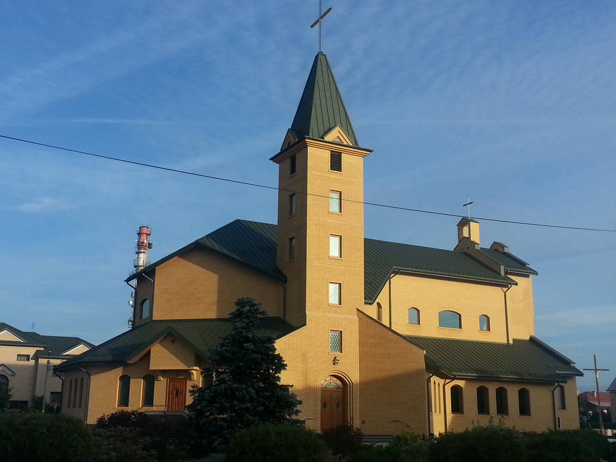 Żółta, murowana świątynia z zielonym dachem i wieżą zwieńczoną krzyżem na tle błękitnego nieba, z widoczną rynną i elementami metalowymi na dachu.