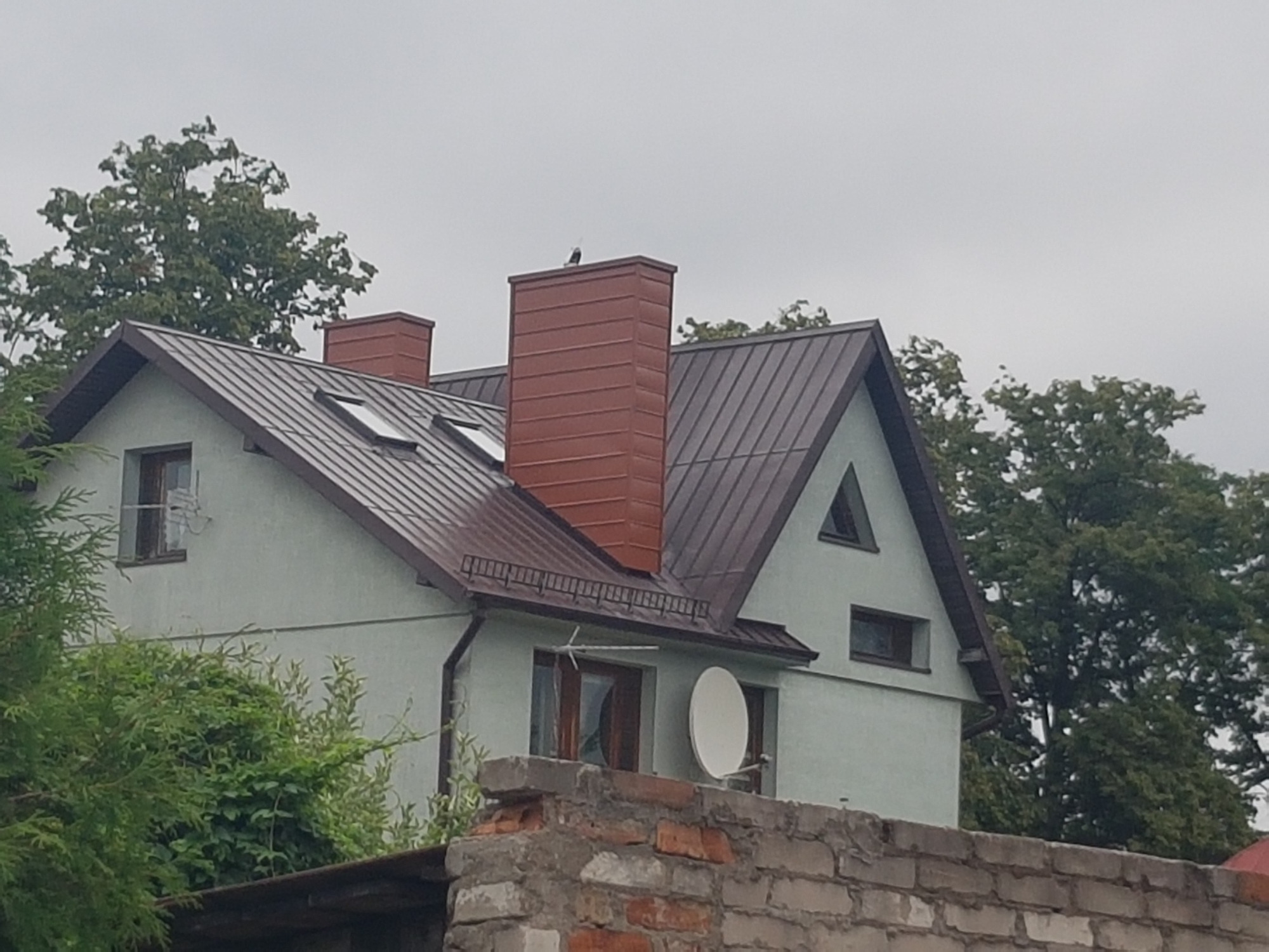 Dom jednorodzinny z nowym, brązowym dachem i kominami w tym samym kolorze, widok zza muru, na kominie siedzi ptak.