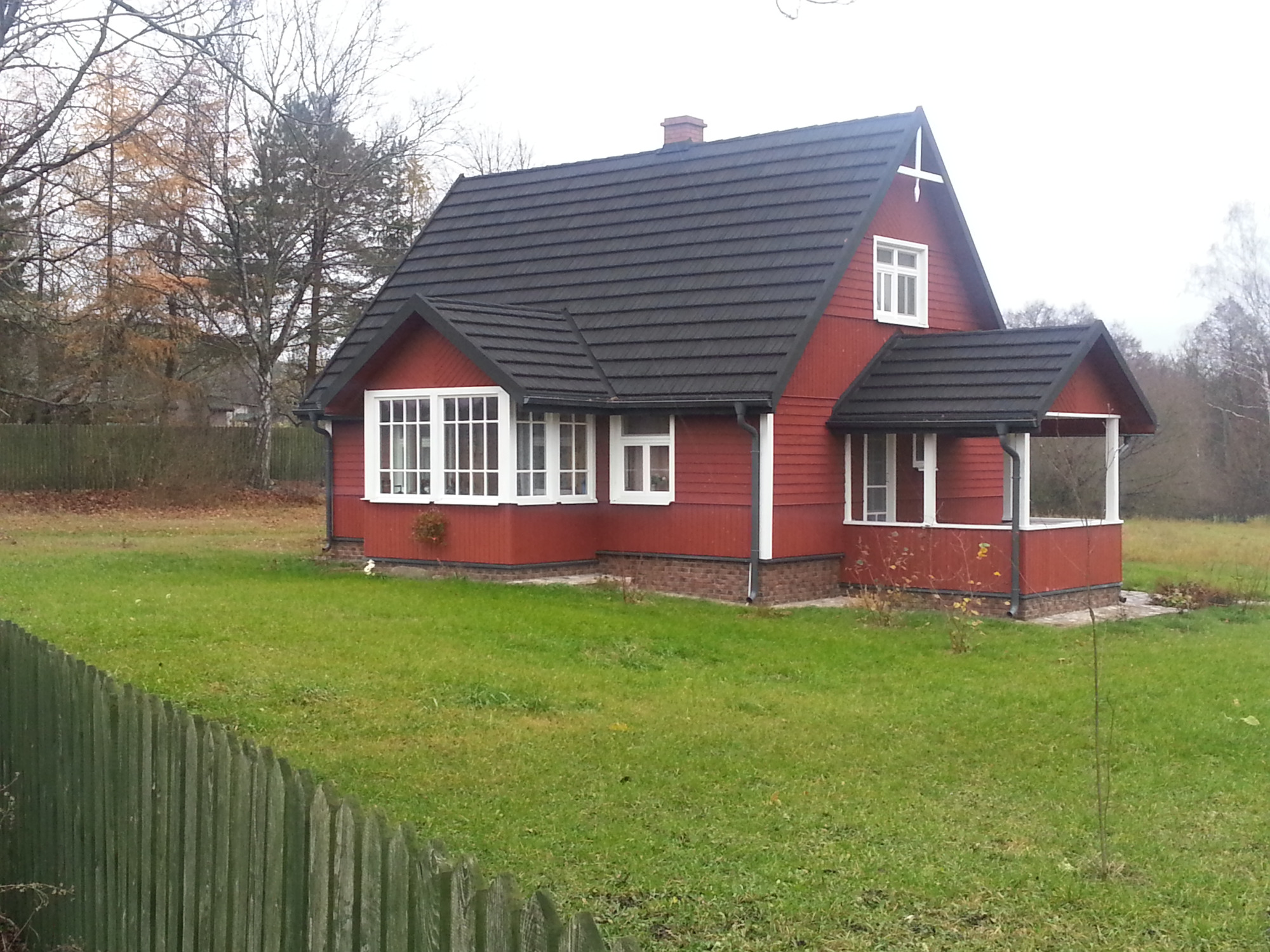 Czerwony dom z ciemną dachówką, widoczny z zewnątrz, z zielonym trawnikiem i drewnianym płotem w tle, w pochmurny dzień.