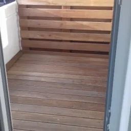 Drewniany taras na balkonie z ażurową ścianką z desek, widok z wnętrza przez otwarte drzwi balkonowe.