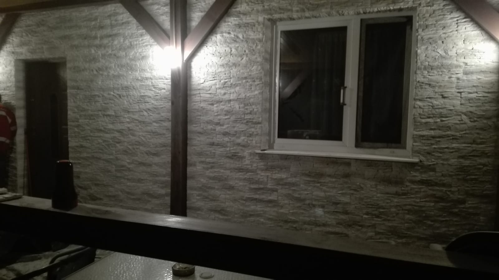 Elewacja budynku wykończona płytkami imitującymi kamień, z oknem PCV i drewnianą konstrukcją, oświetlona sztucznym światłem.