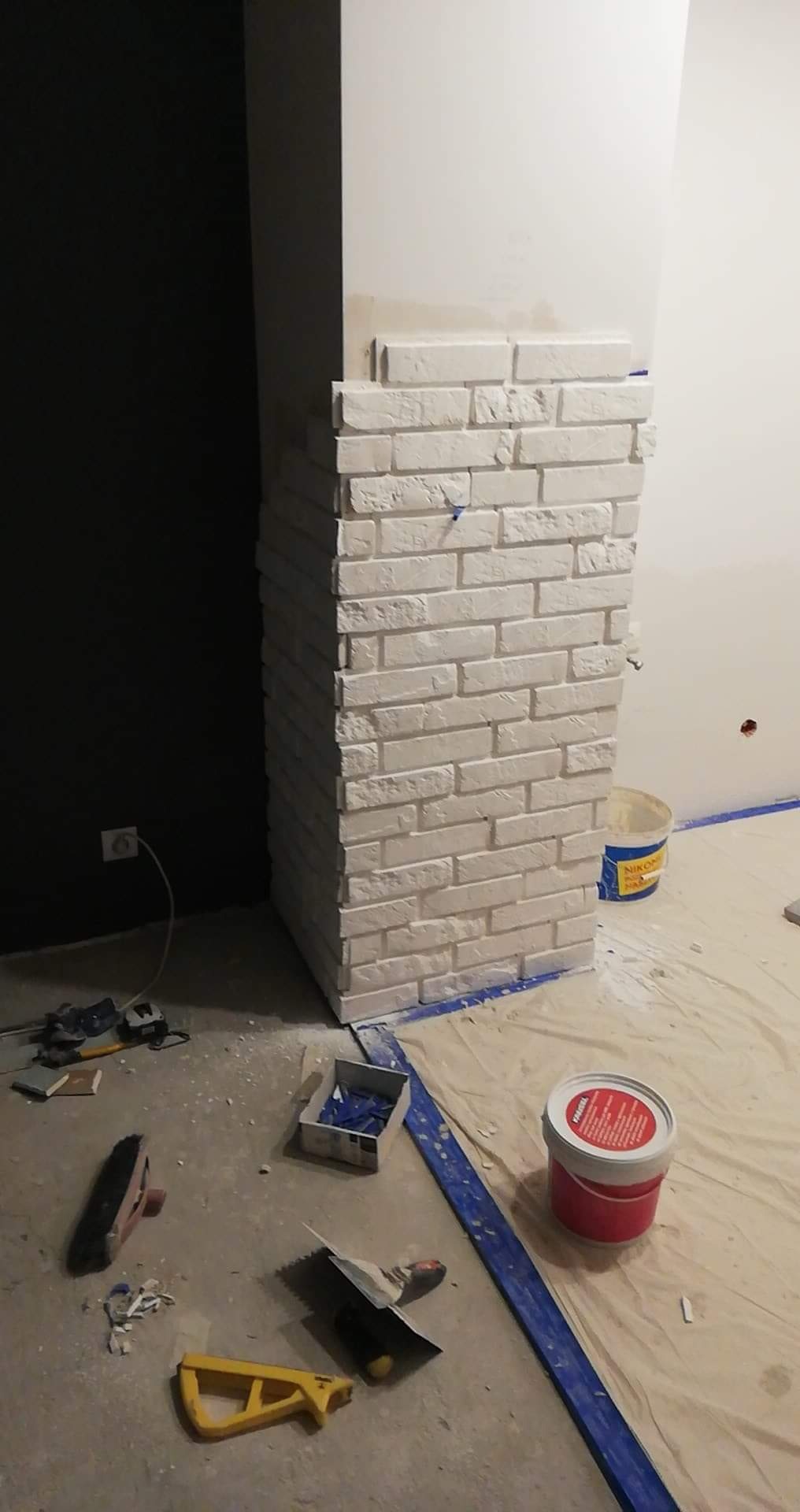 Montaż dekoracyjnej, białej cegły na narożniku ściany wewnętrznej w trakcie prac wykończeniowych, widoczne narzędzia, wiadro z klejem i zabezpieczona folią podłoga.