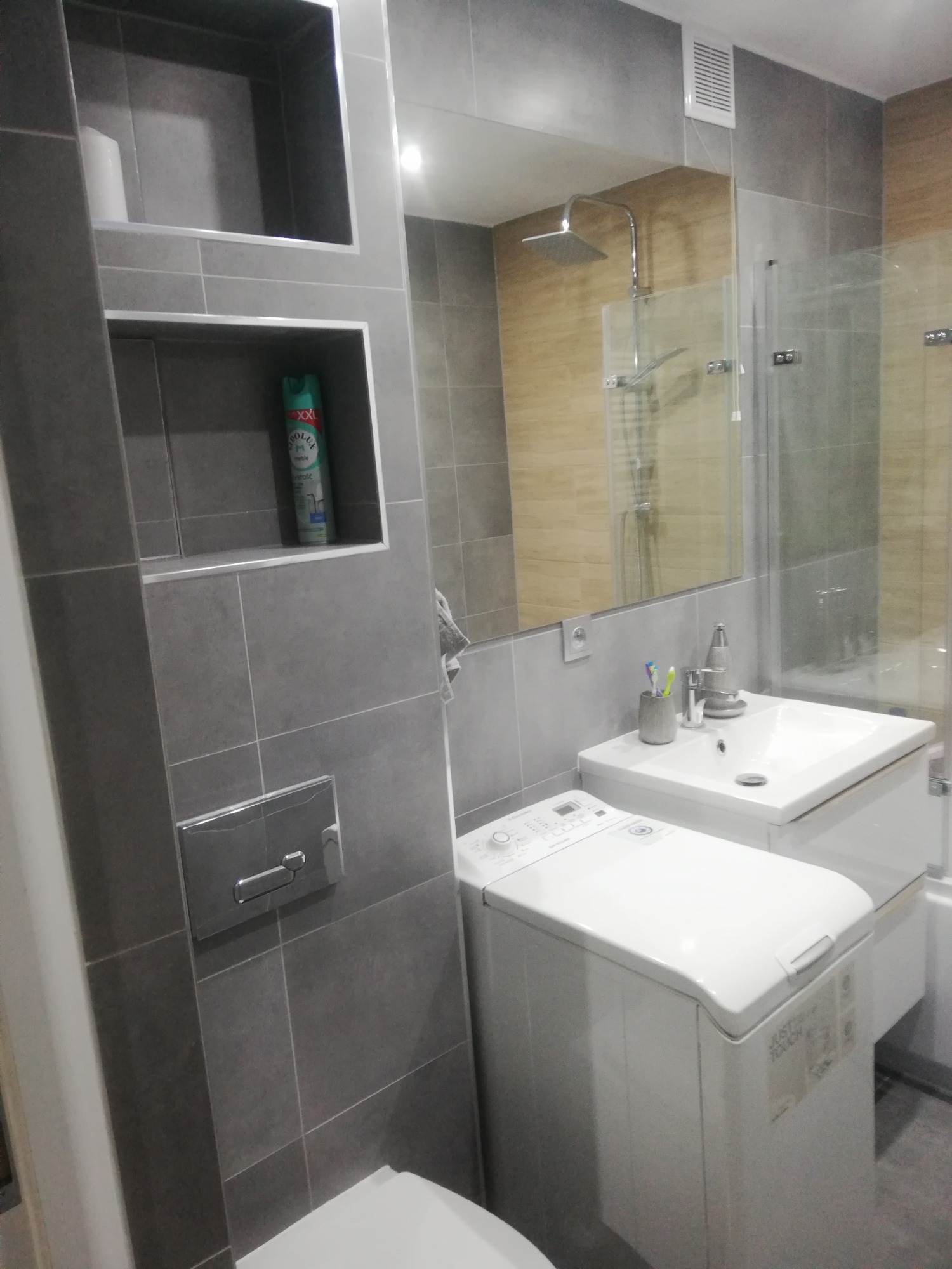 Szara łazienka z wnękami w płytkach, pralką pod umywalką i kabiną prysznicową w lustrze.