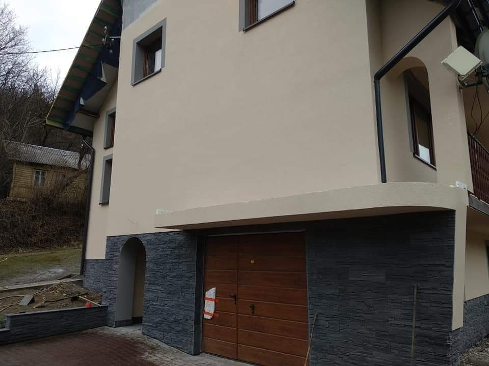 Dwukondygnacyjny dom z beżową elewacją i wykończeniem z szarej imitacji kamienia na parterze, widok na garaż i fragment ogrodu z małym budynkiem w tle.