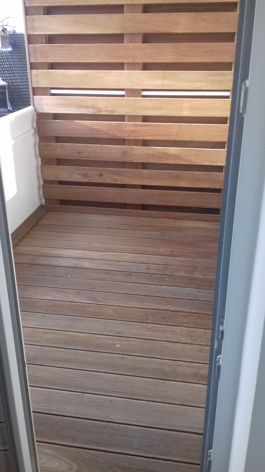 Drewniany taras na balkonie z ażurową ścianką z desek, widok z wnętrza przez otwarte drzwi balkonowe.