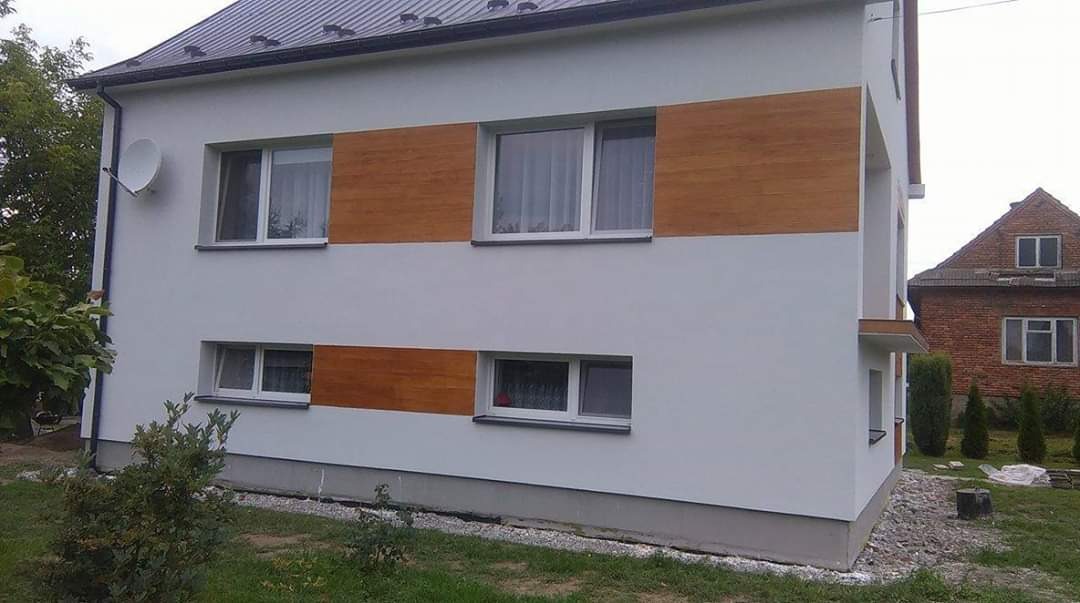 Dwukondygnacyjny dom z białą elewacją i poziomymi pasami z paneli imitujących drewno, widoczna antena satelitarna i sąsiedni dom z czerwonej cegły.