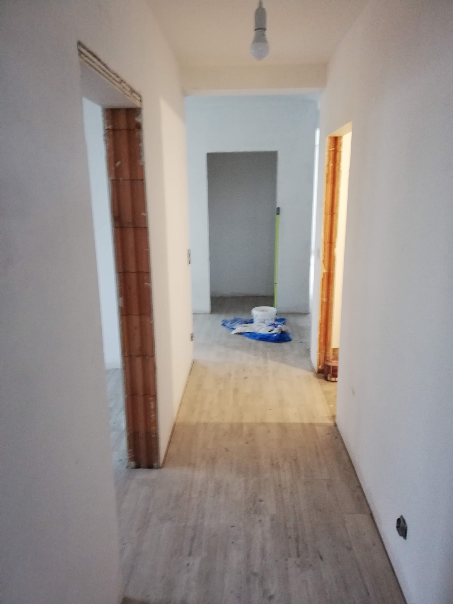 Korytarz w trakcie remontu z widocznymi ościeżnicami drzwiowymi z surowej cegły, świeżo pomalowanymi ścianami na biało i zabezpieczoną folią podłogą.