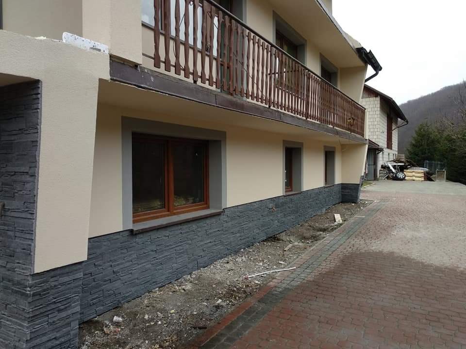 Wykończona elewacja domu z beżowym tynkiem i szarym kamieniem dekoracyjnym na cokole, widok z zewnątrz na częściowo zagospodarowany teren.