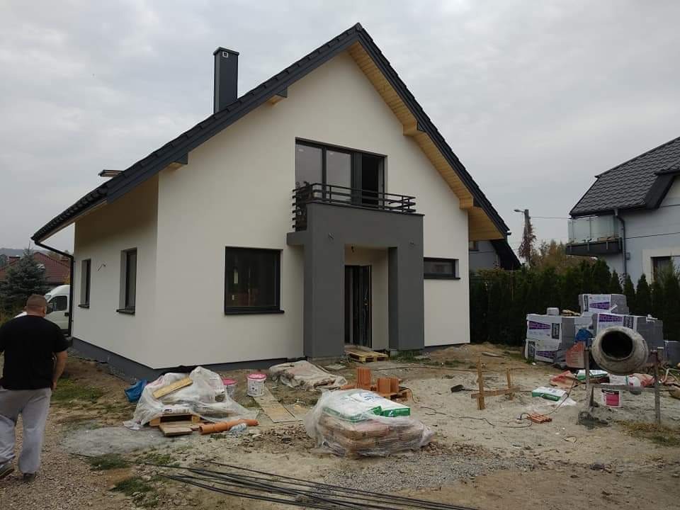 Nowoczesny dom jednorodzinny z ocieploną elewacją w odcieniach bieli i szarości, z widocznym balkonem i elementami wykończeniowymi w trakcie prac. Na pierwszym planie materiały budowlane i betoniarka.