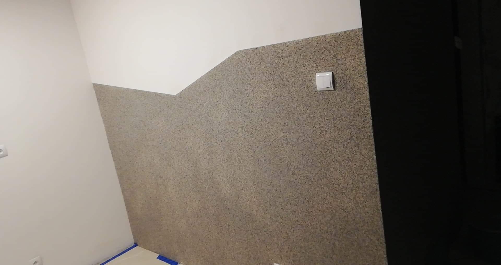 Wykończona ściana z dekoracyjnym tynkiem strukturalnym w odcieniach beżu i brązu, kontrastująca z białą, gładką ścianą powyżej, widoczny włącznik światła i fragment drzwi.