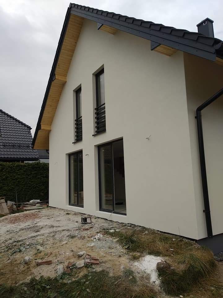 Nowoczesny dom jednorodzinny z jasną elewacją, czarnymi ramami okiennymi i dachem z ciemnej dachówki, widoczne fragmenty niezagospodarowanego terenu wokół budynku.