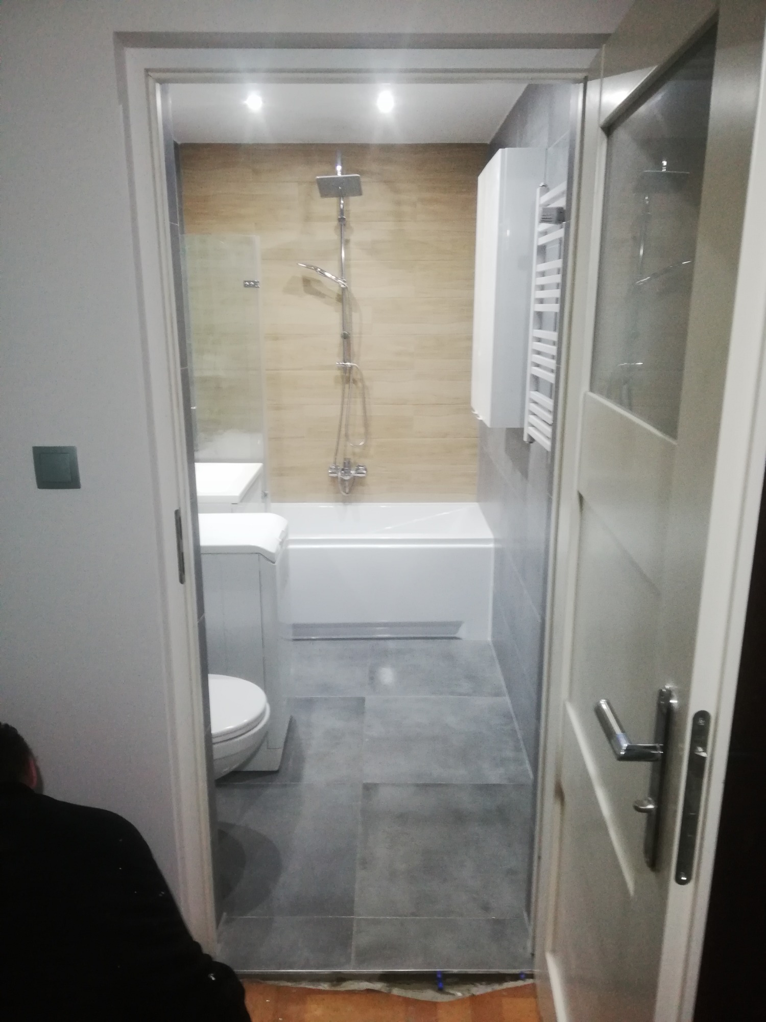 Widok na wyremontowaną łazienkę z kabiną prysznicową, wanną, pralką i grzejnikiem drabinkowym, widzianą zza uchylonych drzwi, z fragmentem osoby w lewym dolnym rogu.