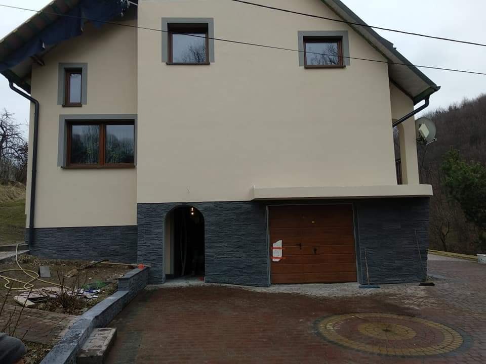 Dom z ocieploną elewacją w odcieniach beżu i szarości, z widocznym fragmentem dachu pokrytego folią ochronną, wskazującym na trwające prace wykończeniowe.