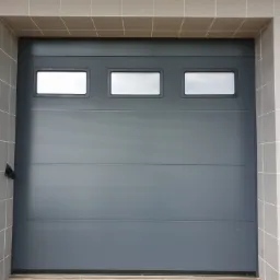 Szara segmentowa brama z trzema prostokątnymi okienkami, zamontowana w elewacji wyłożonej jasnymi kwadratowymi płytkami.