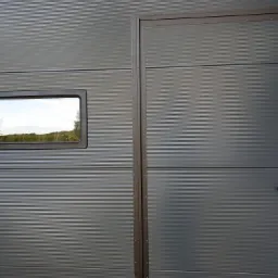 Szara, segmentowa konstrukcja z małym oknem i drzwiami wejściowymi z klamką. Widok na zewnątrz przez okno.