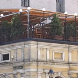 Taras na dachu budynku w stylu kamienicy, z zadaszeniem i oświetleniem, widoczne rośliny w donicach i krzesła.