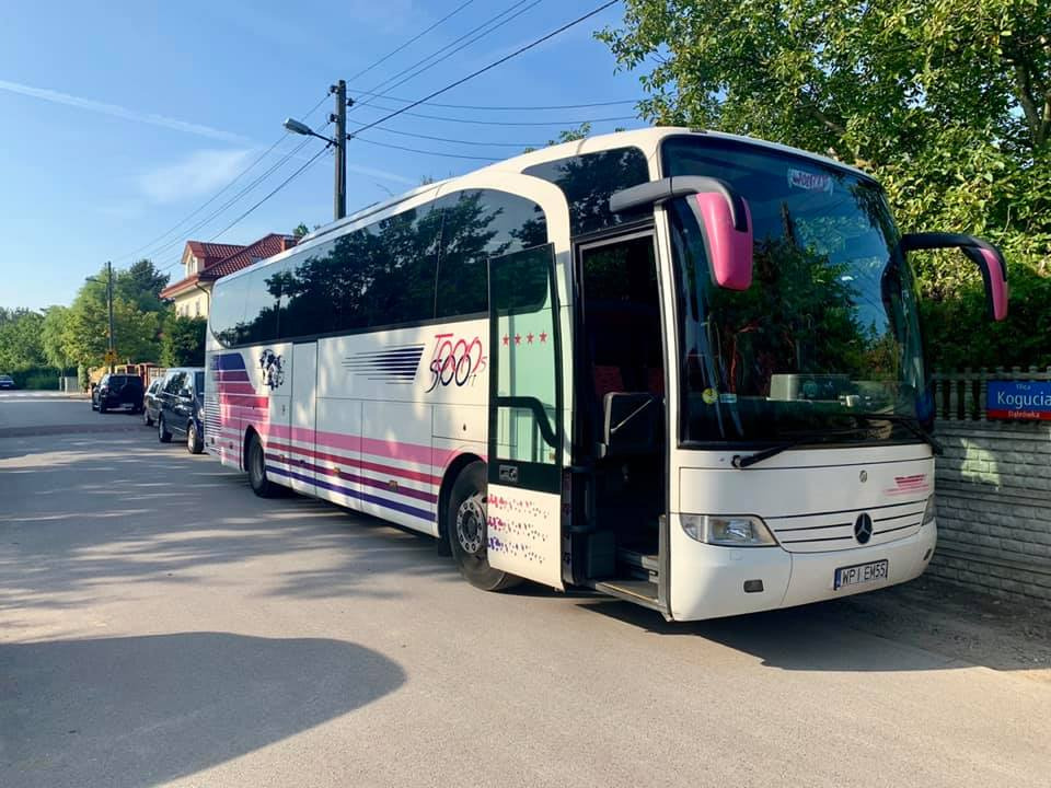 Biały autokar z otwartymi drzwiami i różowymi akcentami, zaparkowany na ulicy z kostki brukowej w słoneczny dzień.