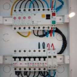 Wewnętrzny widok rozdzielnicy elektrycznej z zainstalowanymi bezpiecznikami i przewodami o różnych kolorach, umieszczonej w białej obudowie.