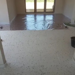 Przygotowanie do montażu ogrzewania podłogowego: siatka grzewcza rozłożona na podkładzie, zabezpieczona folią, widoczne narzędzia i wiadro w rogu pomieszczenia.