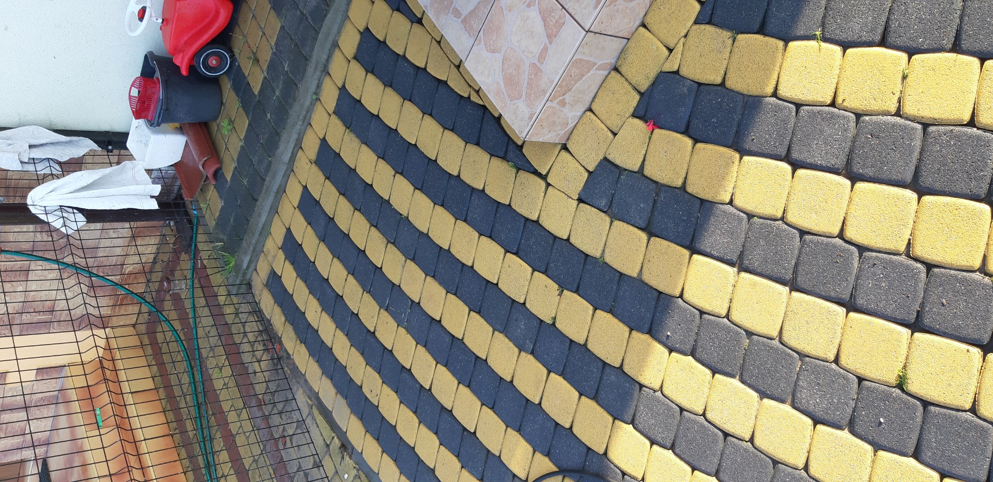 Ułożona kostka brukowa w kolorze żółto-czarnym, ułożona we wzór pasów, z widocznymi drobnymi chwastami wyrastającymi spomiędzy kostek, obok ściany budynku i dziecięcego jeździka.
