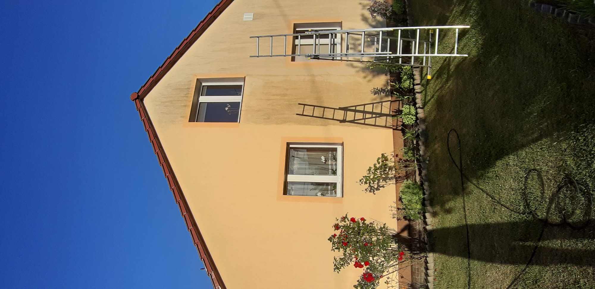 Elewacja domu z widocznymi śladami po czyszczeniu, drabina oparta o ścianę, trawnik z widocznym wężem ogrodowym.