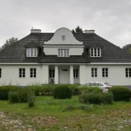 Biały, parterowy dom z dachem z ciemnej dachówki, kolumnami przy wejściu i symetrycznym układem okien, otoczony zielenią i brukowaną ścieżką, z widocznym samochodem osobowym w tle.