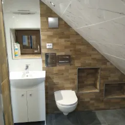 Nowoczesna łazienka na poddaszu w Zbrosławicach z umywalką na szafce, wiszącą toaletą, brązowymi płytkami imitującymi drewno i marmurowymi płytkami na skosach, z wnękami w ścianie.