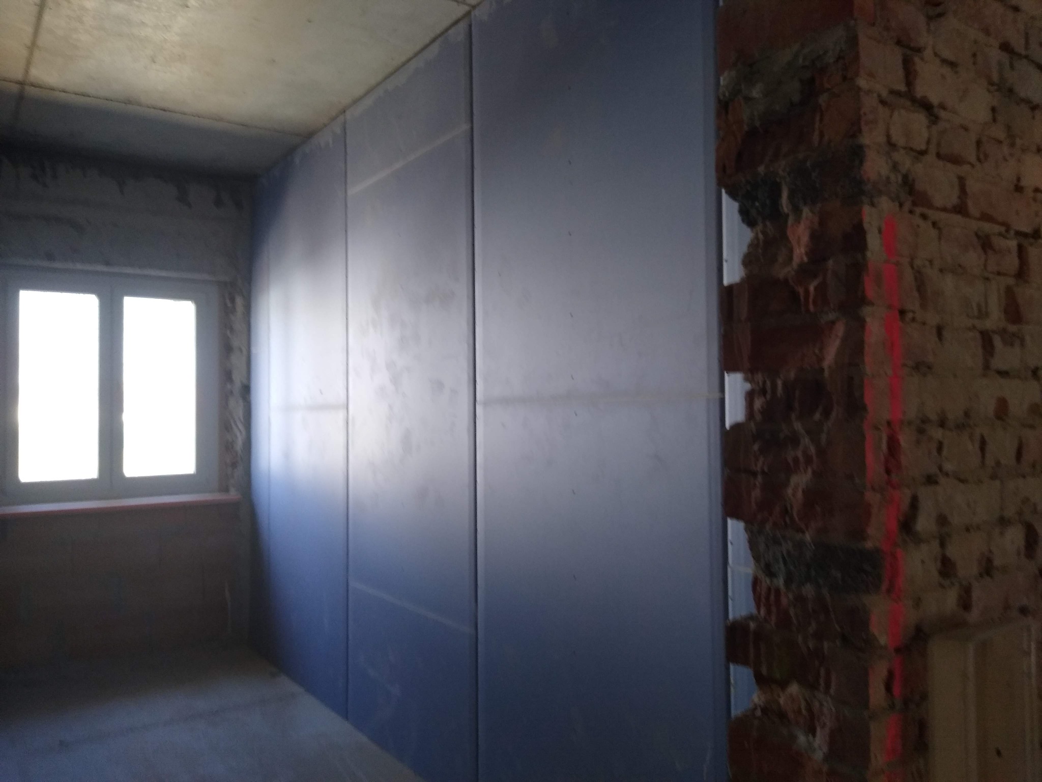 Wnętrze w trakcie remontu: ściana z szarych płyt, fragment odsłoniętej cegły z poziomą linią lasera, okno z białymi ramami.