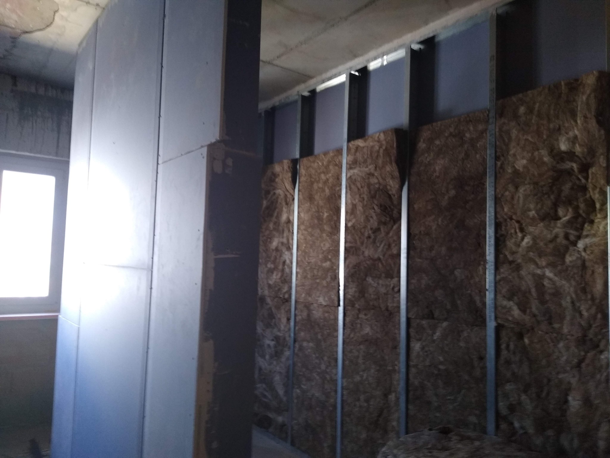 Wnętrze w trakcie prac wykończeniowych: widoczne płyty kartonowo-gipsowe, metalowy stelaż oraz izolacja z wełny mineralnej między profilami.