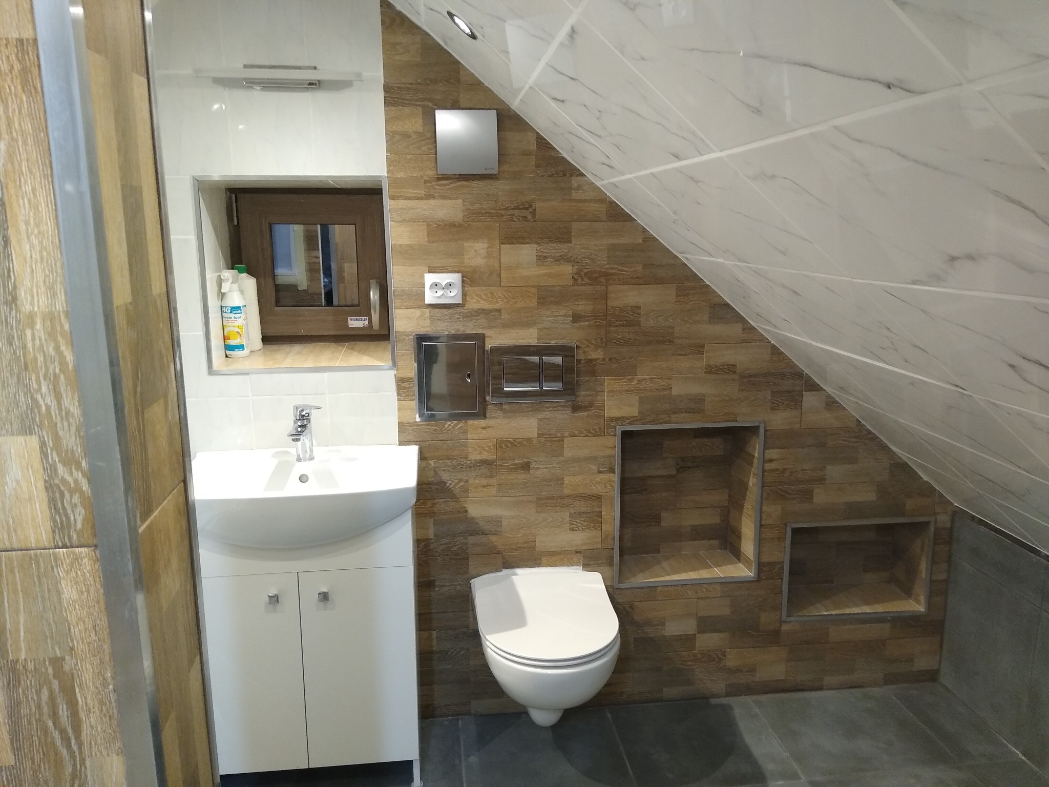 Nowoczesna łazienka na poddaszu w Zbrosławicach z umywalką na szafce, wiszącą toaletą, brązowymi płytkami imitującymi drewno i marmurowymi płytkami na skosach, z wnękami w ścianie.